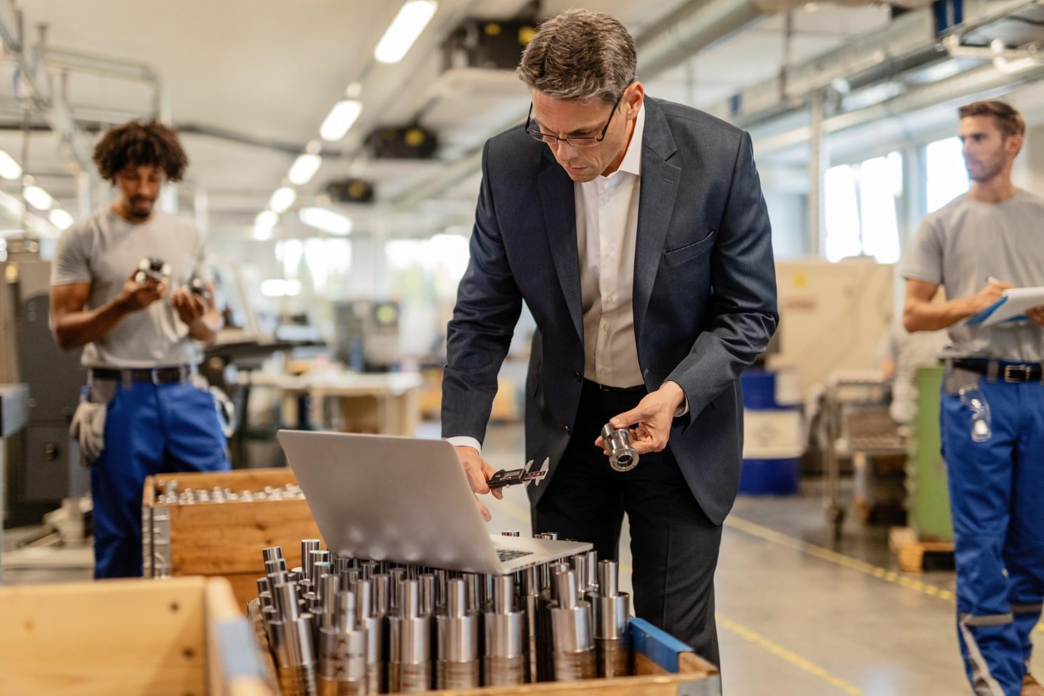 A business professional in a suit carefully inspects a metal component using calipers while referencing data on a laptop in a manufacturing facility.