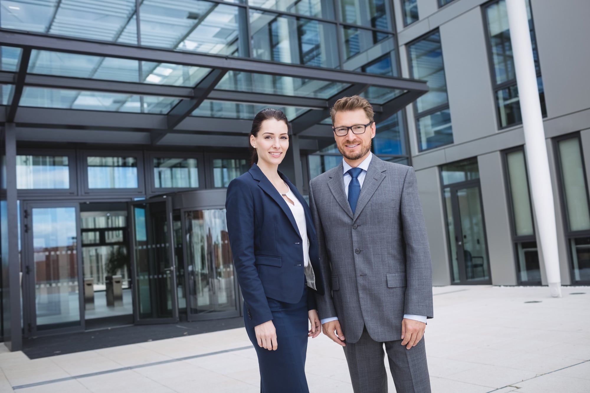 A man and a woman, stand outside a modern office building, dressed in formal business attire.